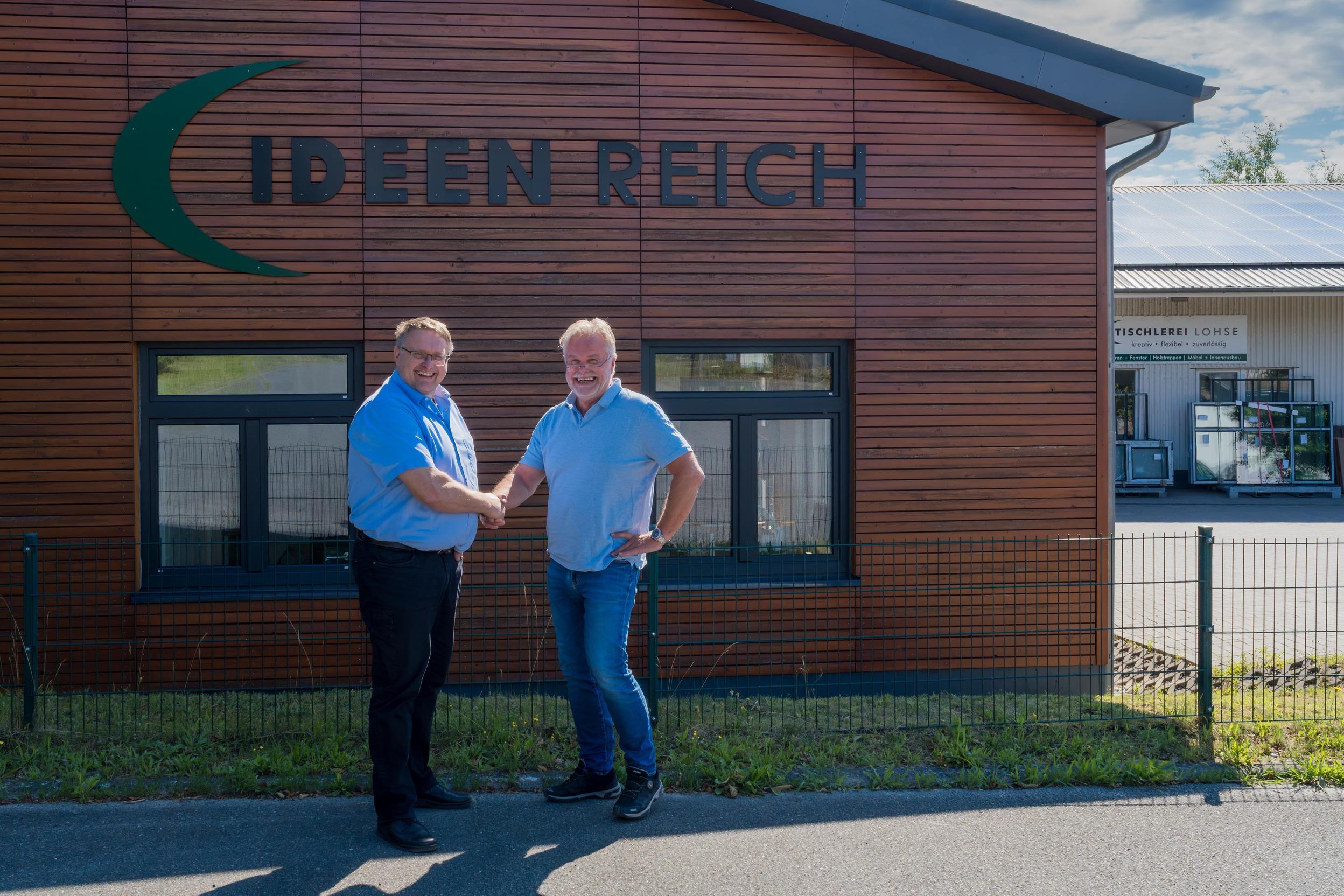 Tischlerei Lohse x Holzträume Lars Lohse und Thomas Jenning in blauen Hemden stehen vor dem IDEEN REICH, welches eine braune Holzfassade hat. Im Hintergrund ist ein Teil der Tischlerei Lohse abgebildet