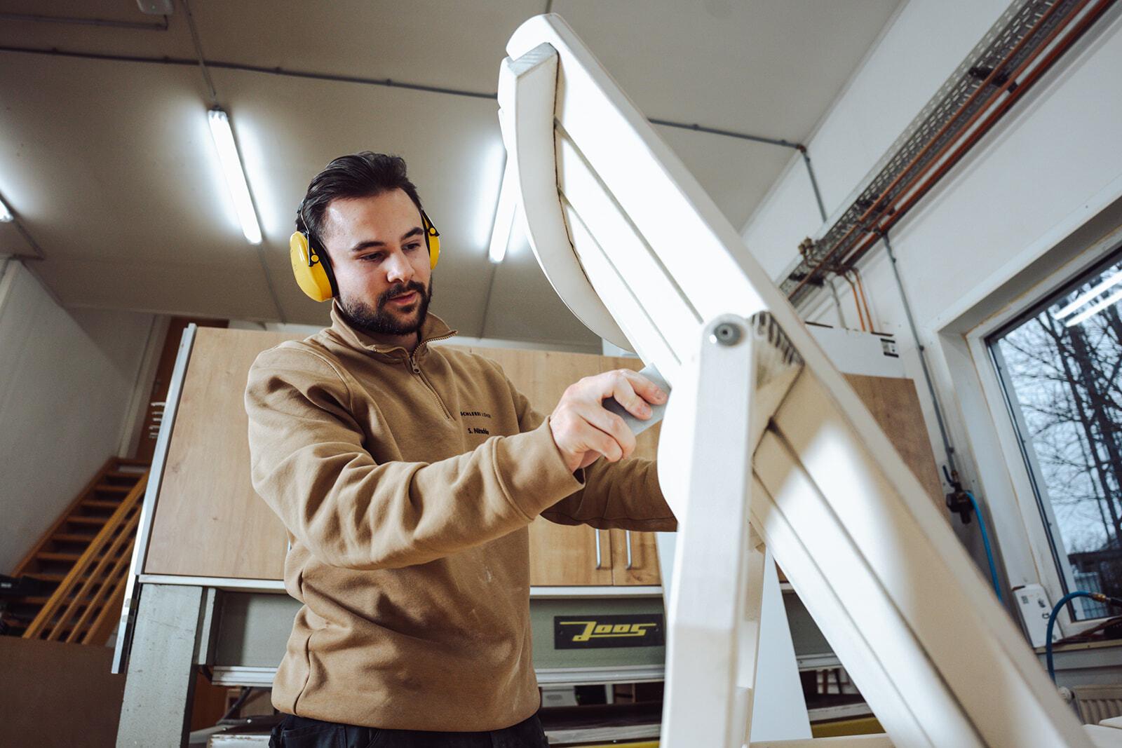 Tischler bei der Bearbeitung eines Holzstuhls Ein Tischler mit Gehörschutz schleift einen weißen Holzstuhl in einer Werkstatt, um eine saubere und präzise Oberfläche zu erzielen.